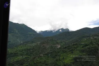 A photograph taken through a vehicle window of steep, forested green mountains under overcast white cloud, with mist hanging over a distant peak. In the foreground hillsides are cut into terraced fields and scattered with small houses and buildings. A dark window frame edge runs down the left side of the frame. No people are visible.