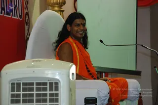 A smiling person in saffron-orange robes with a gold-edged shawl and a dark rudraksha mala sits on a white-draped chair or throne on a stage, with a red banner behind and a microphone on a stand to the right. A white evaporative cooler stands in the foreground. A red dot is on the forehead and long dark hair falls past the shoulders. Background shows a green projection screen and a red banner with partial white lettering.