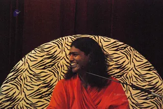 A photograph of a smiling person with long dark hair wearing an orange-red robe, seated in front of a large circular backdrop of cream and black tiger-stripe pattern against dark maroon drapes. A microphone stand angles in from the right. A faint purple bird logo sits in the upper left corner. The person's face is turned to the side, laughing. Appearance-based candidate with long swept hair and saffron-toned robe, but cues are limited, so identity is uncertain.