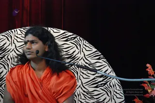 A man with long, swept-back dark hair and a red bindi sits in front of a zebra-striped, rounded chair against a dark red velvet curtain. He wears a loose orange-saffron shawl draped over his shoulders and chest. A gooseneck microphone curves past him toward the right, and orange flowers appear at the lower right edge. The pale chair dominates the frame, and the man looks off to the left. Several cues fit a Swamiji (probable) appearance: long hair, saffron clothing and a sitting position on a raised seat, so he is described here with that label for search.