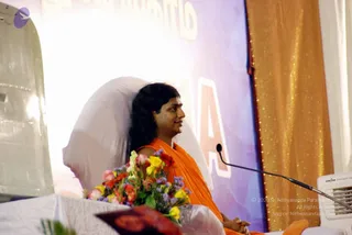 A seated person in an orange saffron robe sits on a stage at a convention, leaning against a large white bolster. Long dark hair falls past the shoulders and the face is turned to the right toward an off-frame point. Hands are folded in the lap. A microphone on a stand stands to the right, and a bouquet of pink, red, yellow and purple flowers rests on the stage in front. A gold shimmering curtain hangs behind, with a purple-and-white backdrop bearing large lettering. Two white air conditioning units flank the stage. Probable Swamiji seated in orange robes with long hair, identified by the page as an event speaker.
