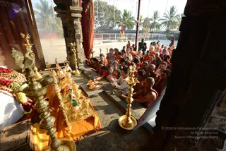 Photograph of a seated congregation in orange and saffron clothes inside a stone temple pavilion at Nithyananda Peetham, Bengaluru. Brass oil lamps stand in the foreground beside a small gold-framed shrine holding a deity murti with a lion mount, on an orange cloth, with flower garlands at left. Rows of devotees, many in saffron robes, sit on the stone floor facing the shrine. One shaved-headed monk in orange sits near the front. A tall palm-lined courtyard and a ratha or chariot are visible outside. The scene appears to be a puja or satsang setting. A figure in dark clothing stands near the courtyard edge at the right, but the appearance is too uncertain to confirm the a person candidate.