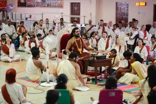 A large hall during a seated Inner Awakening session, with a crowd of white-clad participants wearing red-and-white shawls seated on mats. At the centre, a bearded man with long hair, bare-chested, wearing a rust-red wrap, a heavy gold necklace, a gold cross-shaped pendant and a red-and-gold bracelet, sits on an ornate silver-and-red throne-like chair, gesturing with his right hand. Nearby, a man in white ritual dhoti holds a small ritual object, a woman in a yellow saree sits beside a wooden table with a small drum and a speaker, and two electric fans stand in the foreground. Participants in white and red shawls sit in rows behind, some with white pieces resembling sacred tablets or ritual items on their laps, and a digital clock shows 01:56 in the top right.
