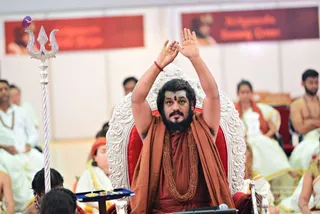 A bearded man in an orange-saffron robe and a long rudraksha mala sits on an ornate silver-and-red throne, raising both hands above his head in a blessing gesture with a red thread on his wrist and a tilak on his forehead. A tall silver trident stands at the left edge. Around him, people in white dhotis and off-white garments sit in rows, some in orange shawls, with a small tray holding a lamp on a blue cloth in front. Red banners with white and gold lettering hang on the wall behind. The appearance-based label is probable; the figure has long swept-back hair, saffron robe and rudraksha mala, matching the page's Swamiji claim. Identity is an appearance-based judgement only.