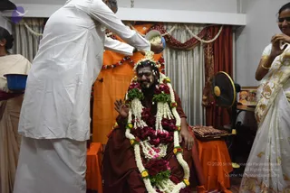 A photograph of a seated man in a maroon-brown robe being honoured with a large garland of white jasmine, red roses and green leaves draped over his shoulders and chest. A man in a white kurta and white dhoti stands behind him pouring white flower petals or rice from a metal bowl onto his head, while a woman in a white saree with gold border on the right smiles and holds her hands together. An orange and red draped backdrop with white flower garlands hangs behind, and a small framed picture stands on the right side. Three people are visible.