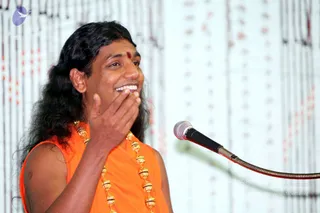 A man with long, dark, wavy hair and a small red bindi on his forehead speaks into a microphone on a stand at right, smiling broadly with one hand raised near his face. He wears a sleeveless saffron-orange garment with a gold-toned ceremonial necklace of large beads. Behind him is a white backdrop hung with vertical strands of red and white beads. a person: long swept hair, saffron clothing and ornate gold necklace, speaking as a discourse presenter on stage.