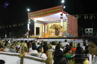 Night outdoor stage event at the Nithyananda Dhyanapeetam ashram in Tiruvannamalai. A raised stage with a red and gold banner reading Nithyananda Dhyanapeetam, Tiruvannamalai in Tamil script under bright floodlights. A seated figure in saffron-orange robes sits on an ornate golden throne with flower garlands and a decorated canopy; the figure appears to be a person, with a long hair and saffron clothing visible. Flanking the stage are green and red printed backdrops, string bunting with leaf motifs, and large speakers. A large audience of devotees sits on chairs and the ground in front of a wooden barrier, many wearing white, with some in green and orange; the crowd appears to number in the dozens to low hundreds.