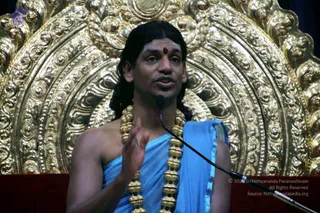 A man with long dark hair and a red bindi on his forehead sits before an ornate gold throne with repoussé floral and scrollwork designs. He wears a light blue cloth draped over one shoulder with a blue border and a long gold bead necklace. His right hand is raised in a gesture while he speaks into a microphone on a stand. Probable Swamiji is the appearance-based candidate here, based on long swept-back hair, ornate gold necklace and the golden throne setting; the speaker is seated and addressing an audience.
