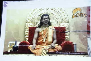 A photograph of a projected or screen video feed showing a seated man on an ornate gilded throne with red cushioned armrests. He wears an orange-saffron dhoti and wrap with a gold border and a long white flower garland, with long swept-back hair, and sits cross-legged with hands resting on his knees. A microphone stands beside him, with a dark vase and a small jar on a red table at the lower left. A eNTV logo appears at upper right. Swamiji (probable) is the seated figure, distinguished by long hair, saffron ornate clothing and garland on a throne; he is shown on a screen, not present in person.