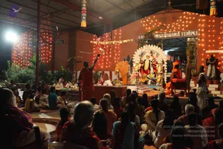 Night photograph of an outdoor temple ceremony in a brick-walled courtyard beneath a corrugated roof, with strings of small lights and a signboard reading Sri Nithyananda Temple. A priest in a red-orange dhoti and shawl stands at the left, raising a lit lamp toward a decorated deity murti flanked by white floral arches. Seated devotees fill the foreground in saffron, pink and white clothing. A figure in orange robes sits on a red-cushioned seat to the right of the shrine, and another person in white stands at far right beside potted plants.