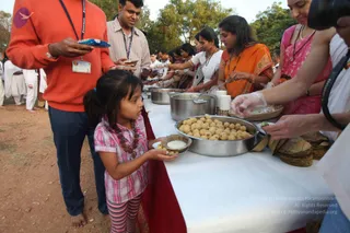 Outdoor food service line on dirt ground beside a long white-covered table with a red cloth underneath. Steel pots and a large steel pan hold round fried balls, with flatbreads stacked at the right and a basket and stack of paper cups nearby. A young girl in a pink plaid shirt and red beaded necklace holds out a small bowl with a white sweet while adults in orange and pink saris and white kurtas serve from the table. A man in an orange sweatshirt with an ID lanyard holds a plate and another man in a striped shirt waits in line. Trees and dusk light fill the background.