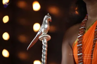 Close-up photo of a silver ceremonial staff topped with a silver parrot figure holding a small red bead in its beak, with a twisted silver shaft. In the right foreground, the shoulder and back of a person in orange saffron robes with a brown rudraksha mala draped over the shoulder, head partly visible at top. Warm blurred bokeh lamp lights fill the dark background. The person's face is not visible.