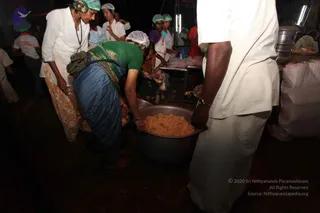 Photograph of a food-preparation scene in a dim tent kitchen during an annadhan-style food service. A woman in a green blouse and blue sari leans over a large black pot of cooked rice, while a woman in a green sari and another in a white top, all with green and white hair nets, stand around steel vessels and serving tables. In the right foreground a man in white shirt and white dhoti stands with his back partly turned. Plastic-wrapped packets and sacks are piled at the right edge.