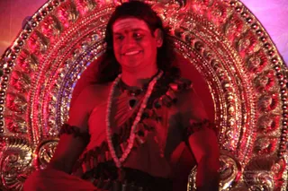 A man sits on an ornate, gilded throne with a large arched back decorated in filigree and beaded patterns, lit with red and magenta stage light. He has long dark hair, a smile, and a painted forehead mark. He wears a red garment with a shawl-like wrap, a rudraksha-style bead necklace, and a dark pendant with layered beads. a person: the seated man has long swept-back hair, a red wrap and beaded adornments on a throne, consistent with the presentation of a person.