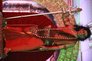 A person in a reclining or lying pose on a red leopard-print cushioned platform, rotated sideways in the frame. The figure wears a flowing red-orange garment, a leopard-print and red bodice, and layered red and white beaded necklaces with rudraksha-style beads. They hold a long silver trident upright in one raised hand, and a small brass vessel is held in the other hand near the platform. Ornate gold-toned gilded arch panels and pink-lit stage backdrops surround the setting. Earrings, bangles and a red forehead mark are visible.