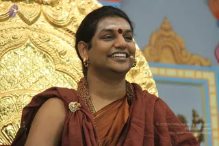 A smiling man with swept-back dark hair wears a red bindi, rudraksha mala necklaces, a gold ornamented brooch and a rust-brown wrap over an orange-saffron garment. He wears a headset microphone and gold earrings. He sits in front of an ornate gold throne with elaborate relief carvings, with a blue and gold framed panel behind him. Appearance-based label: a person, seated as a speaker in satsang.