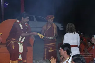 Night-time ritual photograph in a smoky outdoor hall. A man seated on an orange-cushioned throne wears maroon robes, a white and red flower garland, and a tilak, and receives a yellow cloth from a standing attendant in a maroon turban and maroon-and-gold dhoti holding a silver tray. Behind them a white car is parked. At right, a long-haired person in white clothes with a red sash stands facing the scene with hands joined. Seated devotees in orange and saffron robes, some with white headbands, sit along the right side with hands folded in namaskar. A man with red-framed glasses and rudraksha beads sits in front with a microphone. a person is the seated figure, with the throne, maroon robes, and garland as cues. The scene is a ritual or satsang with devotees present.