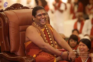 A man seated on an ornate brown leather throne-style chair laughs broadly with eyes closed. He wears an orange-saffron cloth draped over one shoulder and a heavy gold ceremonial necklace with large round gold beads and a rudraksha-style gold chain. A small red dot marks his forehead, and a lapel microphone is clipped near his face. Behind him, several people in orange and white clothing sit in a hall, including a smiling woman in the lower right with a red bindi.