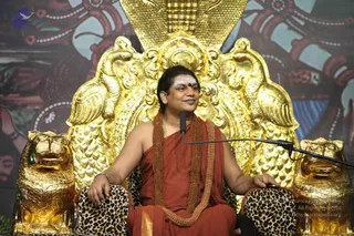 A man seated on an ornate golden throne with a golden hood-like canopy and two golden lion figures at its base, draped in leopard-print cushions. He wears a rust-red saffron robe, a long rudraksha mala and a second mala across his chest, with white vertical tilak and a red bindi on his forehead. He sits with hands resting on the armrests, speaking into a microphone on a stand. Behind him a painted backdrop shows colourful mythological figures.