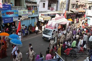 A street procession in a busy Indian town. A white vehicle with a person banner on its roof and a portrait labelled "Paramahamsa Nithyananda" leads through crowded lanes lined with shops, signboards for Reliance Communications and Star Everfresh, and onlookers on balconies. Uniformed police officers in khaki walk alongside, and people in saffron and white stand near the vehicle. Ceremonial umbrellas in orange and blue appear at the left edge, and a red flag hangs from a pole. Pedestrians in saris and kurtas, plus motorcycles, crowd the roadside.