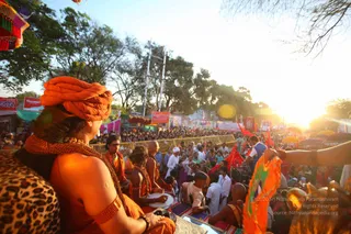 Photograph of a festival procession at dusk, viewed from behind a foreground figure. In the lower left, a sadhu wears an orange turban, orange garments, and a thick orange-beaded mala with a leopard-print shawl over the shoulder, gazing toward the crowd. Beside him, drummers in orange robes play a drum on a raised platform covered with colourful cloth, and bald saffron-robed figures sit nearby. A dense crowd fills the street behind metal barriers, with orange and red flags, banners and umbrellas, and tree-lined sunlit trees beyond. A man in a grey-blue vest stands on the right holding an orange flag. Photo credit overlay and a banner stating '' appear on the image
