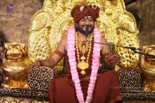 A bearded man in a maroon-red turban with a silver crescent ornament and a red tilak on his forehead sits on an ornate gold throne carved with peacock-like motifs, flanked by gold lion sculptures. He wears a pink flower garland, a heavy gold necklace with a pendant, red beaded armbands and a maroon robe, and smiles while gesturing with his right hand toward a microphone on a stand. Leopard-print cushions cover the seat. Appearance cues (turban, distinctive ornaments, golden throne setting) suggest a person, seated and speaking in a satsang setting.
