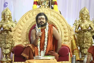 A man seated on an ornate gold throne with red cushioned armrests, wearing an orange-saffron robe and a long white and blue flower garland, holds a microphone and speaks while a steel plate of food sits before him. He has a red tilaka on his forehead and a full beard, and a red thread is visible on his wrist. Golden female deity statues holding oil lamps flank the throne on both sides, and a garlanded kalasha or ritual object appears above. The backdrop is pale lavender draping. A candidate matching the page's cues of a saffron robe, garlands and a throne is present, though the appearance is only partially confirmed, so this is a person in a ceremonial setting.