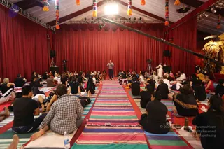 A photograph of a large tented hall with red drapes and hanging colourful paper lanterns, where a crowd sits on colourful striped mats facing a central stage area. Dozens of seated participants wear black and dark clothing, with some in lighter tops. A standing man in a light-coloured shirt and dark trousers stands at the front near the mats, and a figure in white robes stands near the right side beside a golden ornate shrine or deity murti. Speakers, tripods and a boom pole with a camera rig are visible. Water bottles sit among the mats in the foreground.