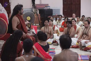 A photograph of a satsang discourse in a hall with cream walls and a wooden door behind. A figure with long dreadlocked hair and a rust-orange shawl over bare shoulders sits at left on a silver-and-red throne, gesturing with one hand toward a standing electric fan. Seated on the floor in front are several devotees: a bare-chested man with rudraksha malas and white dhoti, a woman in white sari with rudraksha, a bare-chested man in white dhoti, and a man in white with rudraksha beads. Others in white and orange saris sit behind, and a tripod-mounted camera and a black speaker stand in the background. Foreground heads are partially visible. A copper vessel sits on the floor and colourful mats are spread out. Visible text reads Vamadeva on a sign above the speaker.