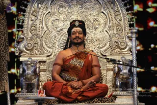 A seated man in an ornate silver throne with elaborate repoussé floral and scroll carvings and silver elephant-head armrests sits cross-legged on a leopard-print cushion before a microphone stand. He wears a red-orange saffron dhoti and a leopard-print shawl over one shoulder, rudraksha armbands, a rudraksha mala, ornate gold necklaces, and a jewelled crown-style headpiece with red trim. A white vibhuti line and red kumkum bindu mark his forehead, and his hands rest in his lap. A small framed photo sits on a stand at the lower left. The backdrop is dark with warm red lighting, and a leopard-print drape hangs at the left edge. a person (a person) is seated as the speaker in this satsang-style setting, matching the page's event context.
