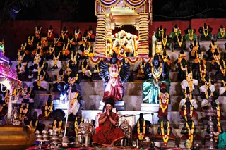 Night-time photograph of a temple ritual before a tiered stone stage lined with rows of seated black-stone murtis dressed in white and red garments and garlanded with yellow and orange marigolds. At the centre top, a multi-armed deity murti stands under a canopy of striped marigold garlands. A man seated cross-legged on a red-brown cloth, wearing a rudraksha mala and red-brown robe with palms pressed together in namaskar, sits at the centre front beside a microphone stand. Brass vessels and a lamp stand sit at the lower left and right, with a decorated palanquin at the left edge.