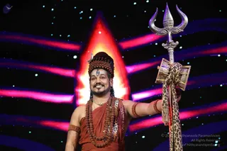 A smiling man stands on a stage against a glowing red-orange flame-shaped backdrop with streaks of blue and magenta light and scattered stars. He wears a rudraksha mala crown with a crescent moon ornament on the headpiece, a white tripundra with a red dot on his forehead, a rust-red dhoti and a leopard-print shawl, plus a rudraksha mala necklace and rudraksha armbands. He raises a silver trident adorned with a leopard-print cloth in his right hand. The appearance of a person with a distinctive rudraksha crown, ornate adornments and trident is consistent with Swamiji (probable), presented here as a Shiva-style stage performer.