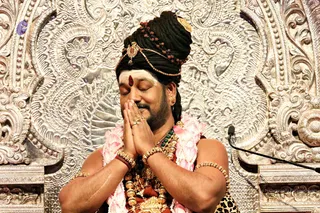 A bare-chested man with eyes closed presses his palms together in anjali mudra before an ornate silver-embossed backdrop, appearing to be at a satsang or puja setting. He wears a tall black hair bun swept upward and wrapped with a crown-like gold ornament and rudraksha malas, a white vertical tilak with a red dot on his forehead, gold earrings, and multiple gold bangles and ceremonial gold necklaces. Pink flower garlands hang over his shoulders and a leopard-print wrap is visible at his waist. A microphone stands at right.
