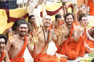 A group of smiling men in saffron-orange dhotis and shawls sits on the floor at a festive meal, many with bare chests and rudraksha malas. Several hold up turmeric-stained palms toward the camera, and a banana leaf with yellow food is spread before them. Behind them a yellow cloth-draped table holds steel vessels, and a few figures in white and orange sit further back. No individual is clearly identifiable as a person by the visible cues, so no candidate is marked.