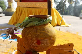 Close-up photograph of a yellow-painted earthen pot (kalasha-style) with a red kumkum mark on its side, wrapped at the rim with fresh green mango or banana leaves. A turmeric root with its hairy roots lies beside it on yellow-painted bricks. Behind it, a golden-yellow draped cloth covers a raised platform, and a colourful rangoli design is visible on the ground. No people are visible in the scene.