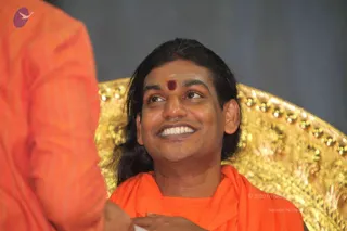 A smiling man with long, swept-back dark hair and a red vermilion dot on his forehead sits in front of an ornate gold throne-style chair with a patterned, sequined backrest. He wears an orange-saffron shawl draped over an orange garment. In the left foreground, a blurred arm in orange fabric reaches toward him. The setting appears to be an event stage with a grey backdrop.