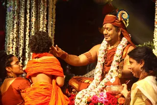 A photograph of a ceremony seated on a decorated dais with white jasmine garlands hanging behind. A man in a rust-red turban with a blue and gold ornament, wearing a red-orange shawl, a gold necklace, a rudraksha mala and a large garland of white and pink flowers, places his right hand on the head of a child in orange clothing who sits with their back to the camera. Two women flank them: one at left in a rust blouse with glasses, one at right in a cream sari holding a bowl. Red roses are arranged at the base of the dais. Swamiji (probable) is the man seated at centre, identified by the turban, saffron-toned robes, garlands and rudraksha cues.