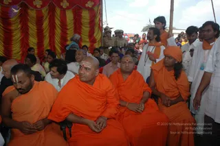 Outdoor gathering in front of a red and yellow draped backdrop with gold flower motifs. In the foreground, several men in saffron-orange robes sit on chairs: a bald man with a red tilak mark sits centre-left, next to another bald man in orange, and a man in an orange turban and orange shawl sits on the right with hands clasped. Behind them stand men in white kurtas, one with an orange shawl and mala, and uniformed police officers stand in the background. Audience members, many women in saris, fill the seats and background, with a red vehicle visible behind. No candidate is confidently identifiable as a person from appearance cues alone, so the figures are described by role.
