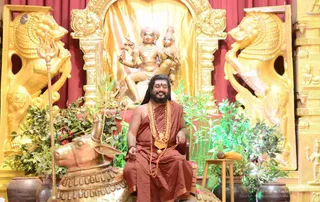 A man with long dark hair and a full beard sits on a golden throne-like seat with a gold-coloured bull statue (nandi) in front of him and a trident leaning beside him. He wears a rust-orange wrap and a heavy gold-toned mala necklace with a rudraksha-style mala draped over his arm, and has a red mark on his forehead. Behind him stands a golden deity murti of a seated figure with a companion, framed by gold lion-like carved panels and red drapes. Potted green plants, a small wooden stool with a coconut and fruit surround the seat. The setting is a golden stage at a satsang-style gathering.