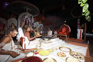 A night-time outdoor ritual under a canopy, with a white square fire pit or homa altar at centre surrounded by brass plates of flowers, sweets, red powder and grains. Several white-clad priests sit on the floor; the foreground priest has white-painted forehead markings and gold chains. Behind them, illuminated white and decorated deity murti and figures stand on the left. On the right, a seated figure in saffron-orange robes and a beaded necklace sits on an ornate red and gold throne, with a standing attendant in white and orange-red shawl nearby. Strings of lights and tassels decorate the tent.