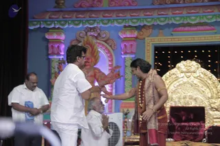 A photograph on a stage set with a painted blue and pink backdrop and a colourful deity figure. A man in a white shirt and white trousers, seen from behind, bends forward with his palms pressed together and reaches toward a man in saffron-and-maroon robes wearing a shawl, rudraksha-style beads and gold bangles. The robed man, with long swept-back dark hair, extends his hand in greeting beside a large gold-carved throne with a red cushion. A seated elderly man in white folds his hands in namaskar at lower left, and another man in white stands at left with his hands clasped. Warm stage lights glow in the background.