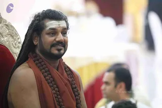 A photograph of a bearded man with long dreadlocked hair and a white tripundra mark on his forehead, seated near an ornate gold-and-red chair. He wears an orange-red shawl over bare shoulders and a long mala of dark brown rudraksha beads. He smiles gently while looking off to the side. In the blurred background, another man in white clothing sits near a red and gold setting. Overlay text reads , , Source