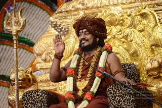 A man seated on an ornate gilded throne with leopard-print cushioned armrests, smiling and raising his right hand in greeting while speaking into a microphone on a stand. He wears a maroon-brown turban with a gold crescent ornament, a rudraksha mala and a rudraksha bracelet, a long white and orange-red flower garland, a gold pendant necklace and a saffron-brown robe. A golden trident stands to his left beside a gold lion-head armrest, and the background shows marigold and white garlands framing the stage. Probable Swamiji seated in a ceremonial setting, with cues of maroon turban, rudraksha mala and ceremonial gold necklace.