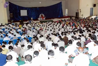 Wide view of a large hall with a blue draped backdrop and a central stage. Hundreds of seated devotees fill the floor, most in white clothing with white headbands, sitting on green mats and blue chairs. On the stage a figure in orange-red clothing sits on a white throne-like seat, with a projection screen to the left. A crowd of people in white with white headbands fills the foreground.