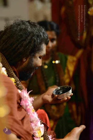 A close profile photograph of a bearded man with long dreadlocked hair wearing an orange-brown sannyasi robe and a long garland of pink and white flowers with yellow accents, a rudraksha-style mala visible at the chest. He leans forward looking down into a small ornate metal bowl held in his open palm, which holds a dark liquid. A blurred figure behind him in a green garment with a beaded mala and gold-toned ornaments is also present, with red and gold drapery in the background. The setting appears to be a ritual or satsang gathering.