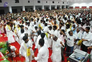 A large hall crowded with devotees in white dhotis and white shirts, many with yellow cloth bags, raising hands in a devotional gathering. A red carpet runs along the front, with a tripod, potted plants and a projector on a table in the lower corner. A man in an orange shirt stands among the crowd, and the back wall shows red-trimmed windows and ceiling fans. The scene appears to be a convention session, with people visibly numbering in the hundreds.
