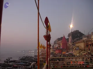 Dusk view over the Ganges at a Varanasi ghat, with a row of moored wooden boats along the left riverbank and the still, hazy water beyond. Red metal railings and red poles with small yellow flags and red lamp holders line a raised walkway on the right. A tall red temple spire rises among older multi-storey buildings along the bank, a street lamp glows above, and small groups of people stand along the ghat in the fading light.
