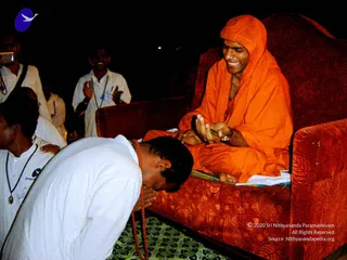 A photograph at night of a smiling person seated on a red upholstered sofa, wearing a saffron-orange robe with the hood drawn over the head and a wristwatch on the left wrist, holding a small object in cupped hands. A man in a white shirt bows forward with his head lowered, a red-orange rudraksha-style mala hanging from his neck. Behind them, two men in white shirts with lanyards stand with palms pressed together, and a partly visible man on the left holds a camera. The seated figure is an appearance-based a person, as the page indicates, with saffron robe and hooded head as the distinguishing cues; the bowing devotee and standing attendees are in white. A green patterned mat lies on the floor.