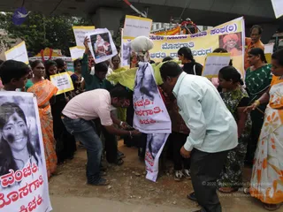 A street protest under a concrete overpass with a crowd of men and women holding yellow-bordered placards and a large yellow banner. A man in a pink-and-white striped shirt and jeans bends over a cloth effigy printed with a face and Kannada lettering, while a man in a light blue shirt leans in beside it. Women in orange, green and floral saris stand around, and one holds up a printed photo of a woman in red and white. A banner shows a portrait of a saffron-turbaned man labelled in Kannada. Several protesters bend and crowd close to the effigy on a dirt road.