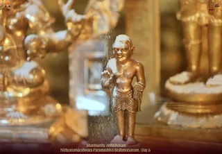Close photograph of a small golden metal statue of a bare-chested, dhoti-clad figure standing in the centre, its head and shoulders dusted with white powder and a fine spray of liquid or powder falling around it during abhishekam. Ornate gilded deity figures flank it on the left and right, also dusted white, and the caption reads Shatmurthy Abhishekam, Nithyanandeshwara Paramashiva Brahmotsavam - Day 6. No physical people are visible; the figure is a statue.